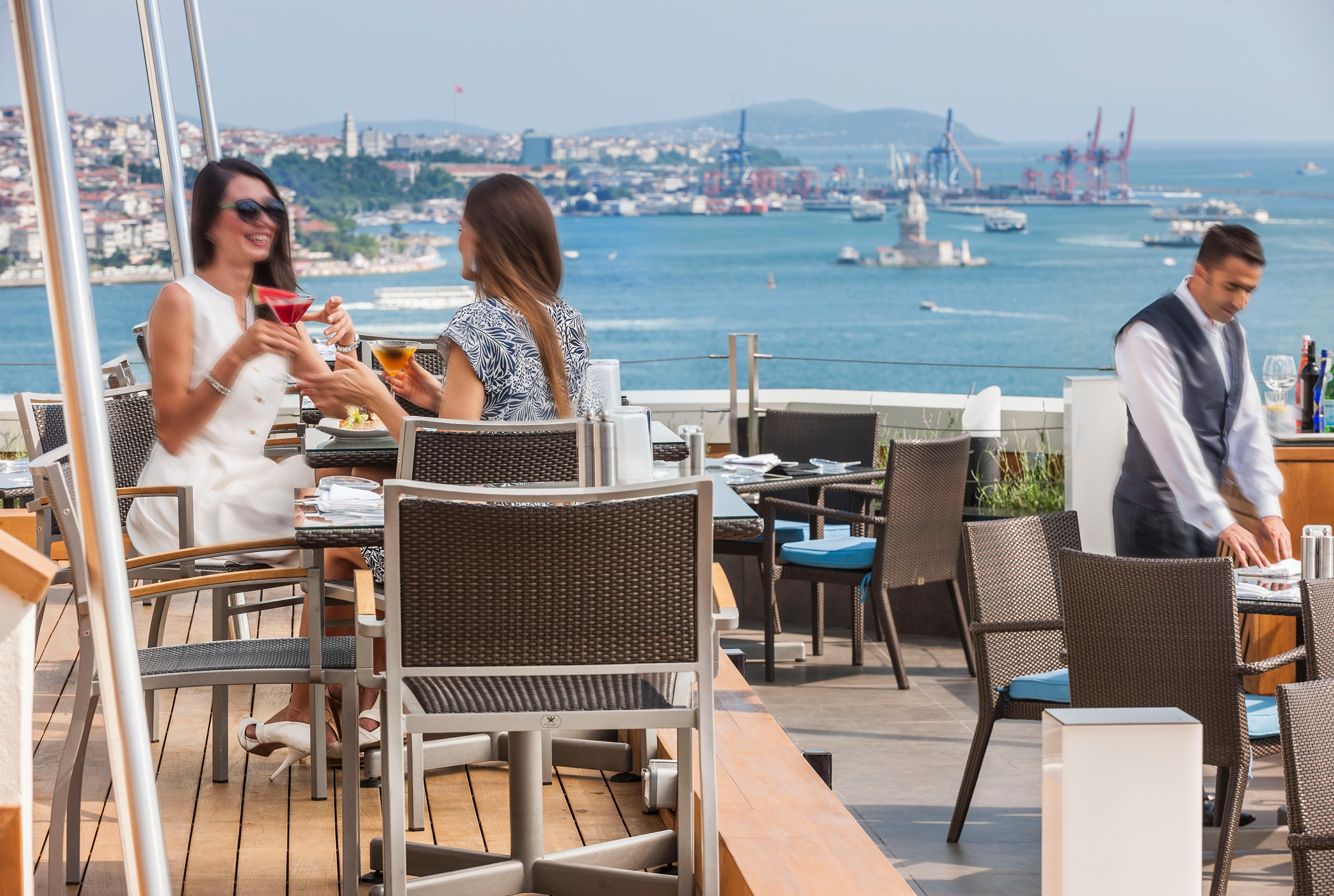 Swissotel The Bosphorus Istanbul Luaran gambar The photo depicts a vibrant outdoor setting at a restaurant with a view of the sea and distant cityscape. Two women are seated at a table, enjoying drinks and engaged in conversation, while a waiter in a uniform is nearby. The environment appears rel