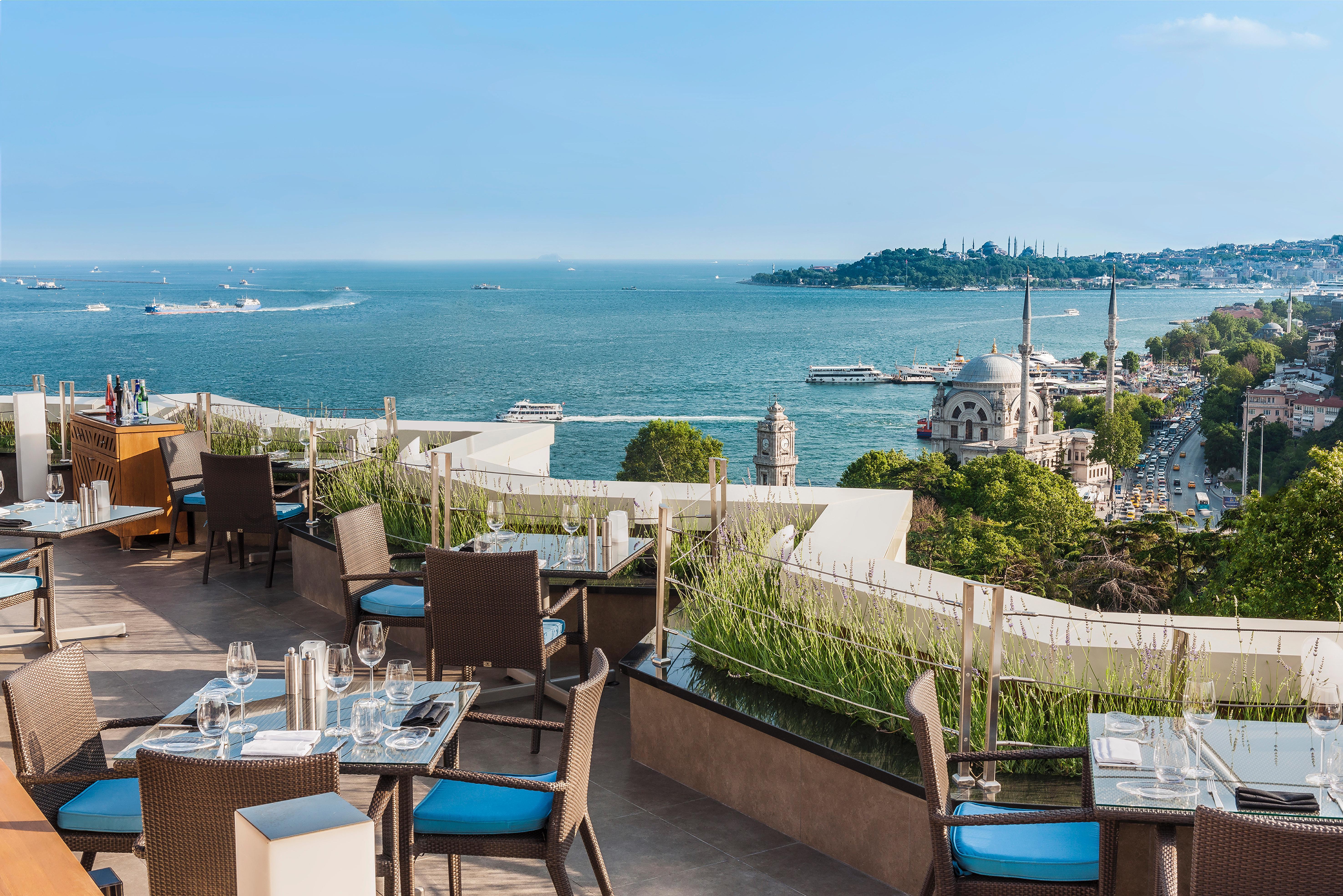 Swissotel The Bosphorus Istanbul Luaran gambar The image shows a beautiful outdoor dining area on a terrace overlooking a scenic coastal view. The setup includes several tables with elegant dining arrangements, featuring chairs with blue cushions. In the background, there is a vast body of water,