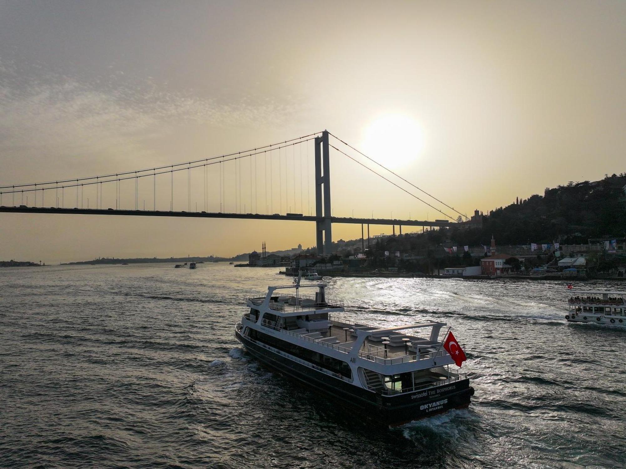Swissotel The Bosphorus Istanbul Luaran gambar The photo depicts a scenic view of a large body of water with a ferry boat traveling across it. The ferry is adorned with a Turkish flag. In the background, there is a bridge spanning the water, with the sun setting or rising behind it, casting a war