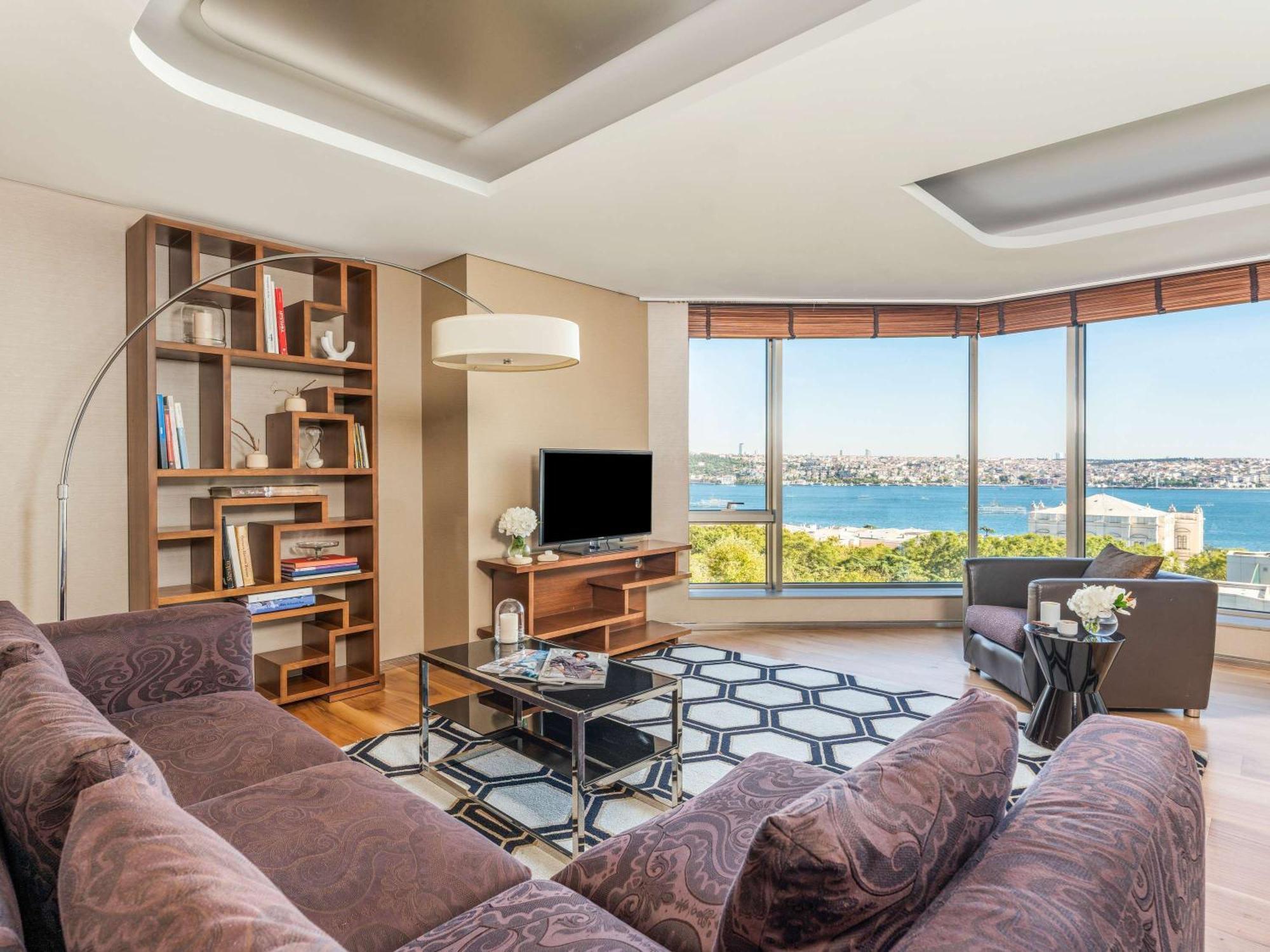 Swissotel The Bosphorus Istanbul Luaran gambar The photo shows a modern living room with a large window offering a scenic view of a body of water. The room features a brown sectional sofa, a glass coffee table, and a stylish rug with a geometric pattern. There is a contemporary floor lamp and a b