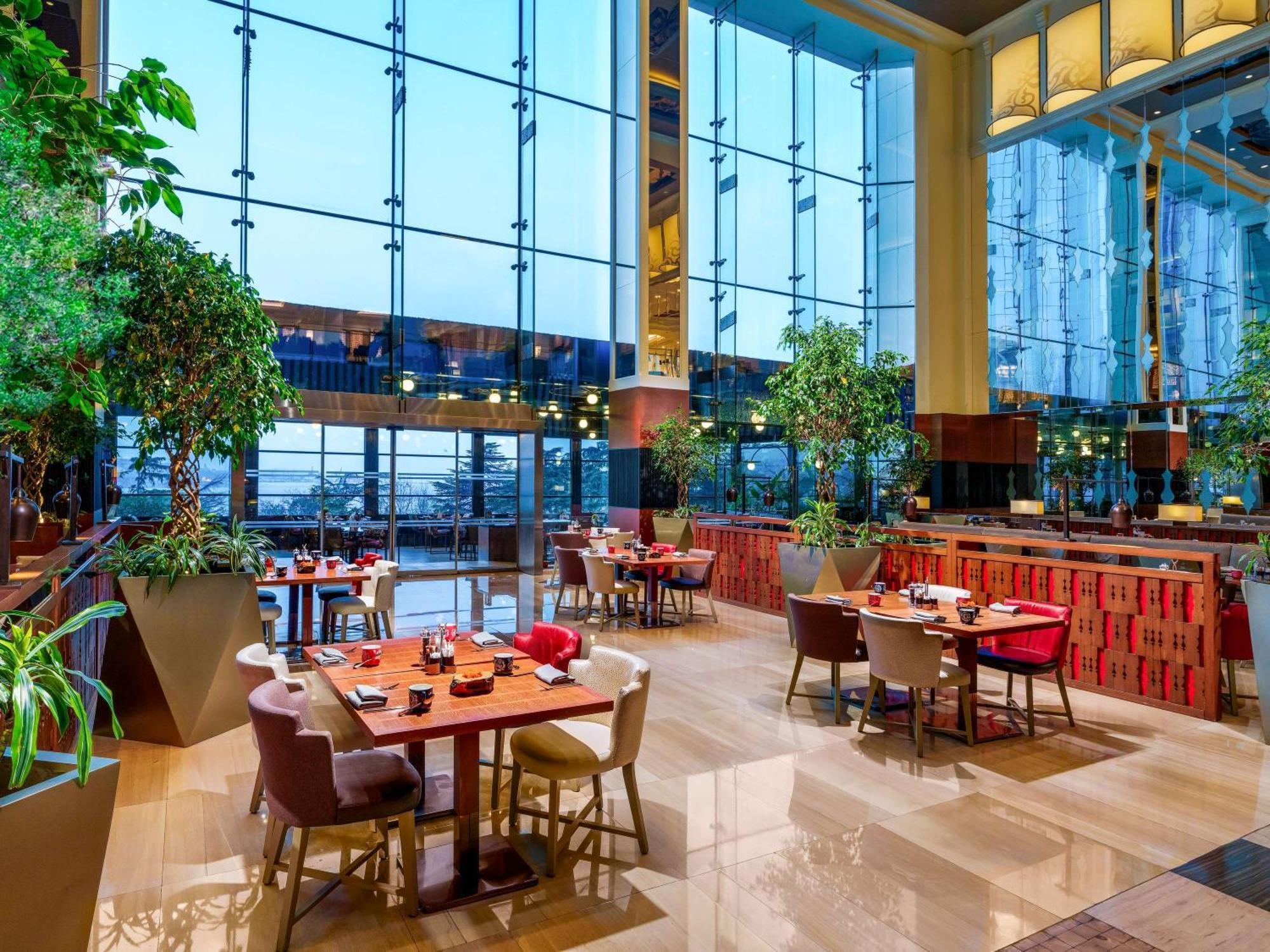 Swissotel The Bosphorus Istanbul Luaran gambar The photo shows a modern restaurant interior featuring large glass windows that allow plenty of natural light to enter. The decor includes lush greenery, with potted plants placed throughout the space. There are several dining tables arranged comfort